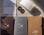 A styled flat-lay of stationery and packaging for 'the off hours', including a white envelope, two round black-and-white stickers, a printed card with a minimalist beach scene, and three rectangular paper goods in dark brown, charcoal gray, and kraft brown with white typography.