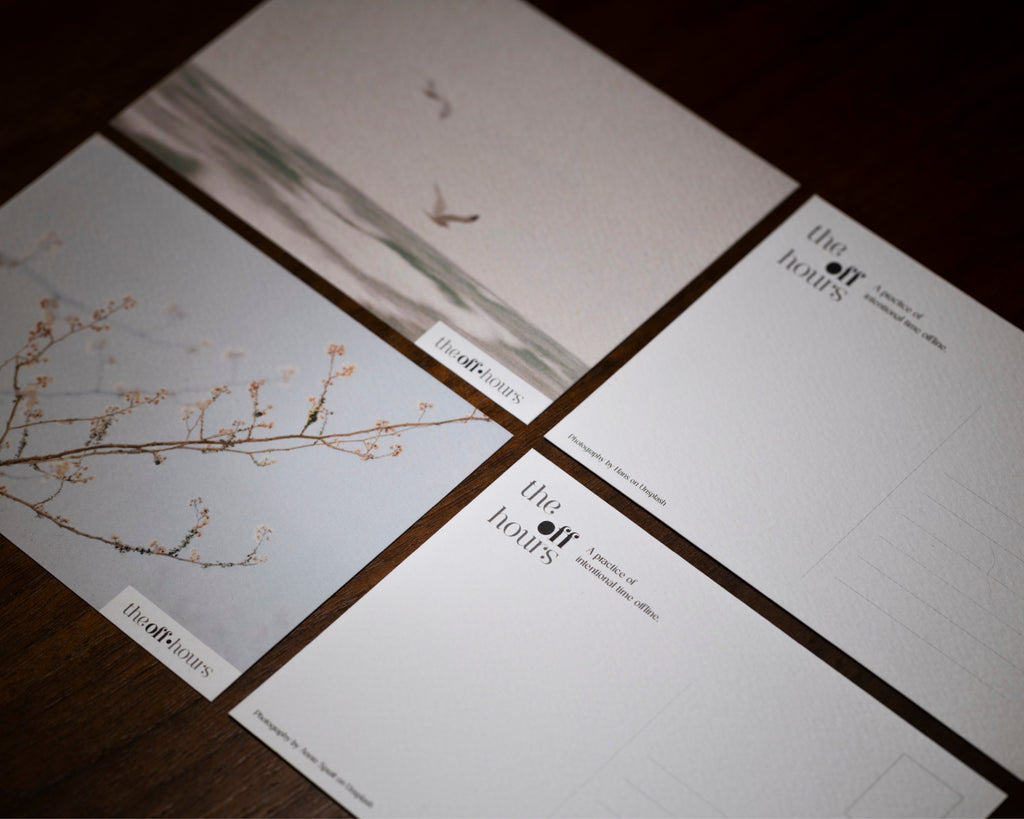 Four printed cards or postcards arranged in a grid on a dark wooden surface. The cards feature minimalist artwork, including a bird over water and delicate blossoms, with the text 'the art books' and smaller copy printed on them.