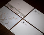 Four printed cards or postcards arranged in a grid on a dark wooden surface. The cards feature minimalist artwork, including a bird over water and delicate blossoms, with the text 'the art books' and smaller copy printed on them.